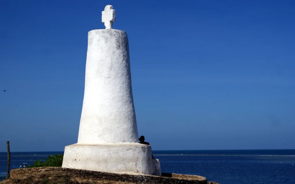 A tall, slender white stone pillar topped with a small cross, standing on a reinforced concrete base against a bright blue ocean backdrop.